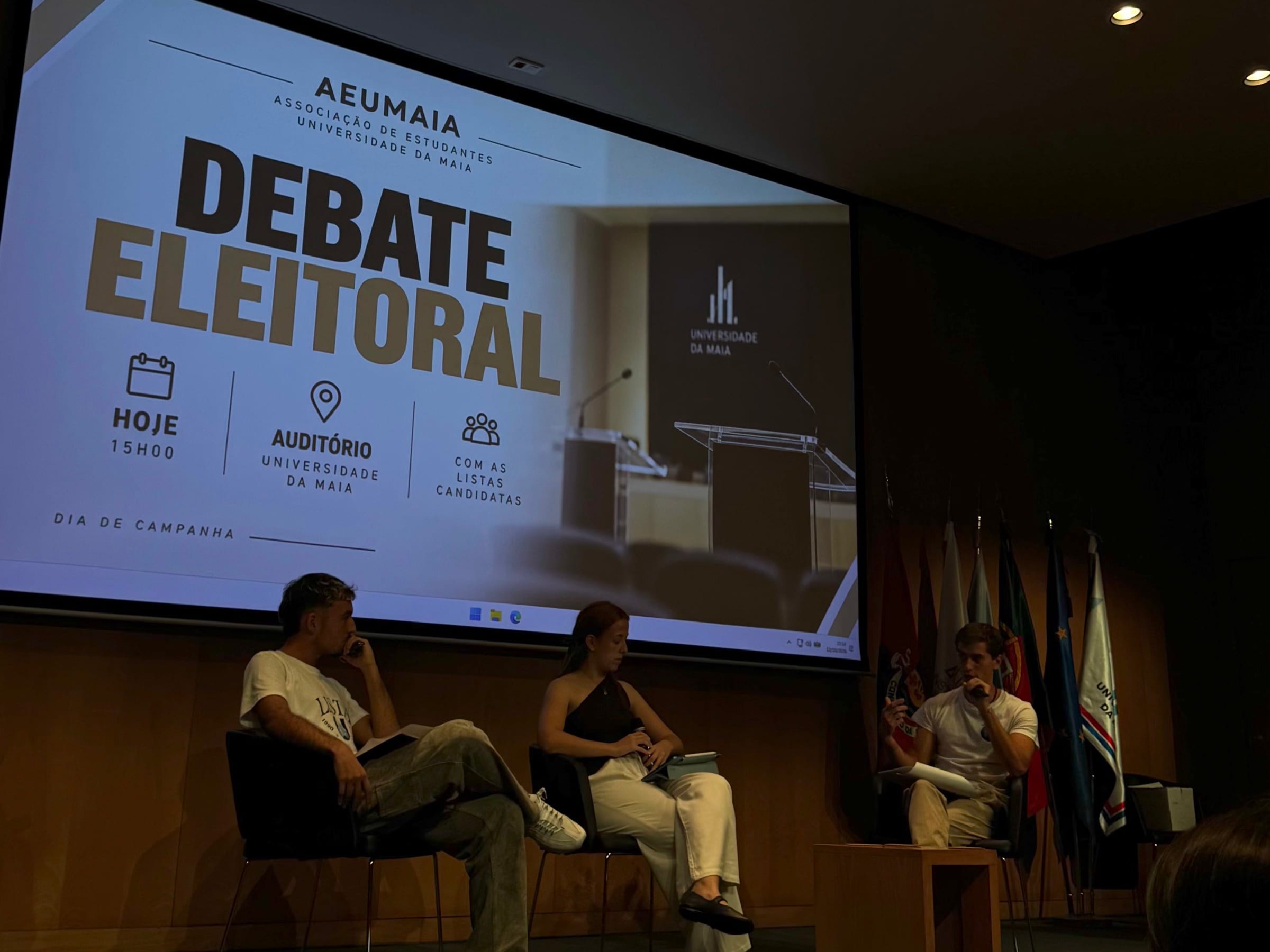 AEUMAIA election debate in the University of Maia auditorium, with three people seated on stage in front of the debate screen